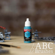 Bottle of the Army Painter Fanatics in the colour Phalanx Blue with various wargaming models and an ABC paintbrush in the background, on a wooden surface.