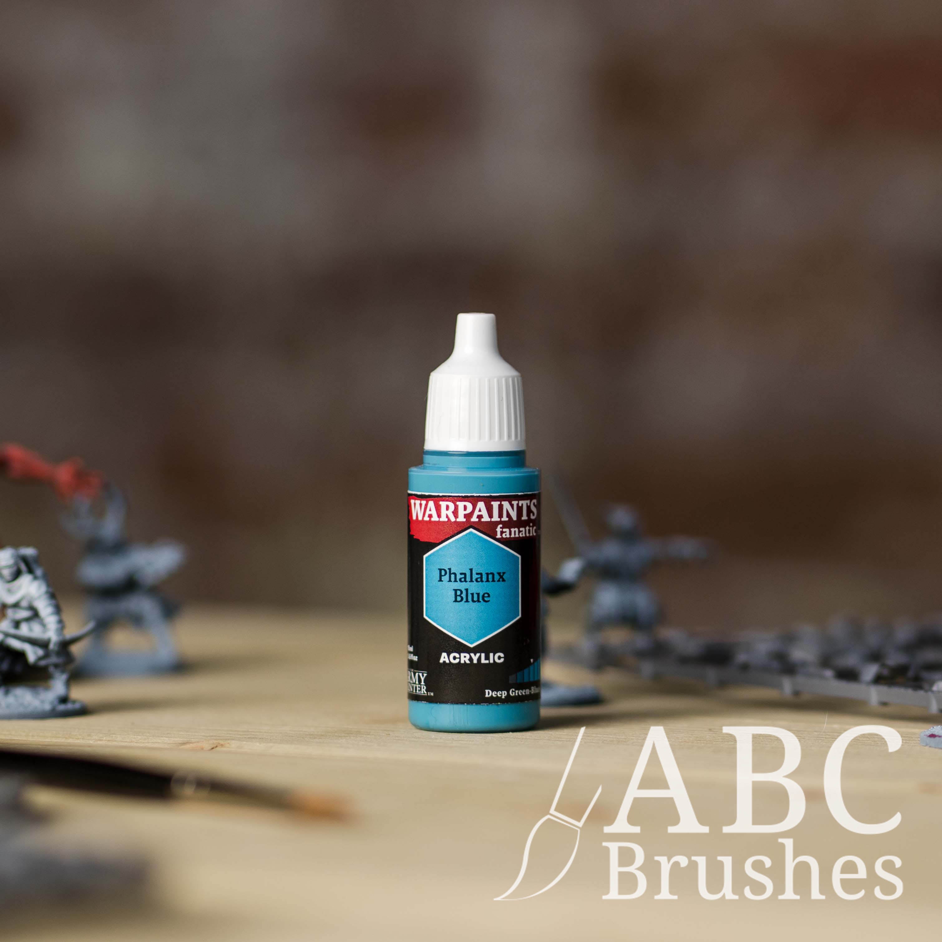 Bottle of the Army Painter Fanatics in the colour Phalanx Blue with various wargaming models and an ABC paintbrush in the background, on a wooden surface.