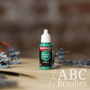 Bottle of the Army Painter Fanatics in the colour Pharaoh Guard with various wargaming models and an ABC paintbrush in the background, on a wooden surface.