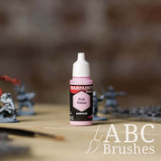 Bottle of the Army Painter Fanatics in the colour Pink Potion with various wargaming models and an ABC paintbrush in the background, on a wooden surface.