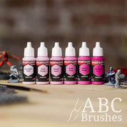 Army Painter Fanatics Pinks Triad on a wooden surface. There are wargaming models and an ABC paint brush next to them.