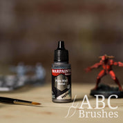 Bottle of the Army Painter Fanatics Metallic paint in the colour Plate Mail Metal with various wargaming models and an ABC paintbrush in the background, on a wooden surface.