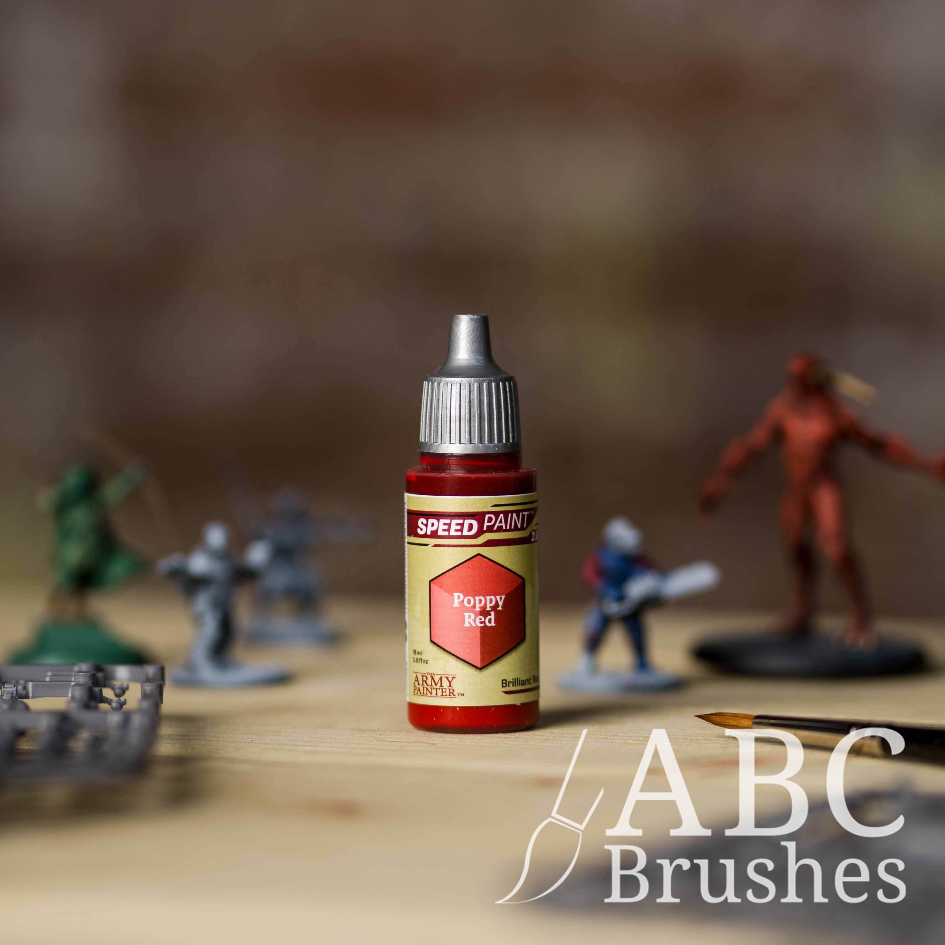 Bottle of the Army Painters Speedpaint in the colour Poppy Red, with wargaming models and an ABC paintbrush in the background, on a wooden surface.