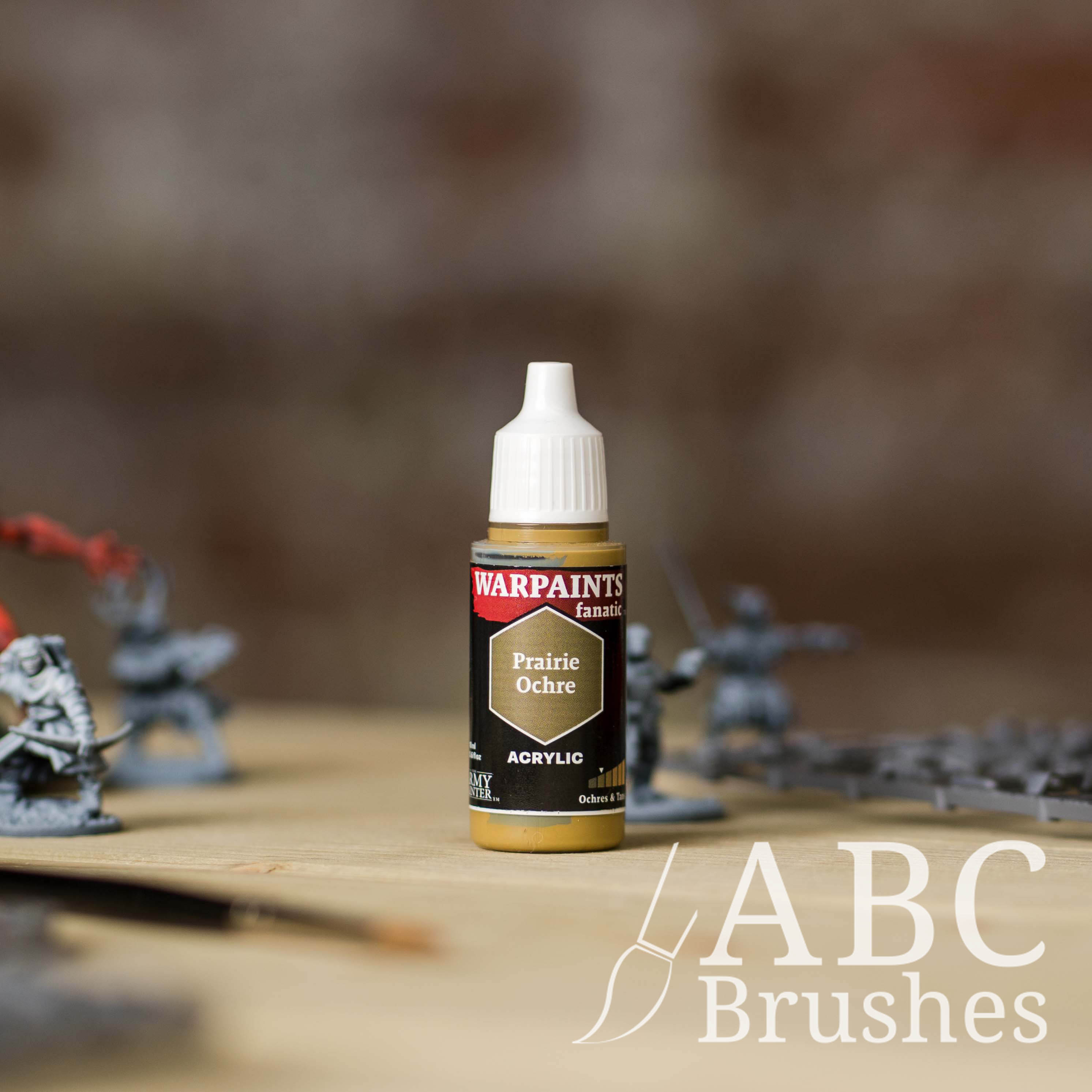 Bottle of the Army Painter Fanatics in the colour Prairie Ochre with various wargaming models and an ABC paintbrush in the background, on a wooden surface.