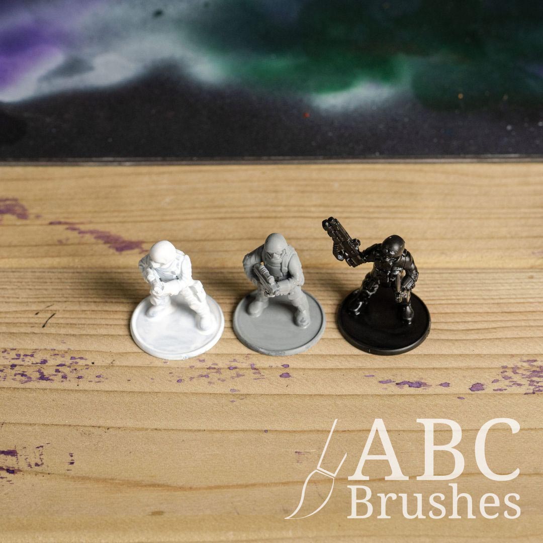 Three soldier miniatures with guns in various poses, on a wooden surface. From Left to right, they are painted white, grey and black.
