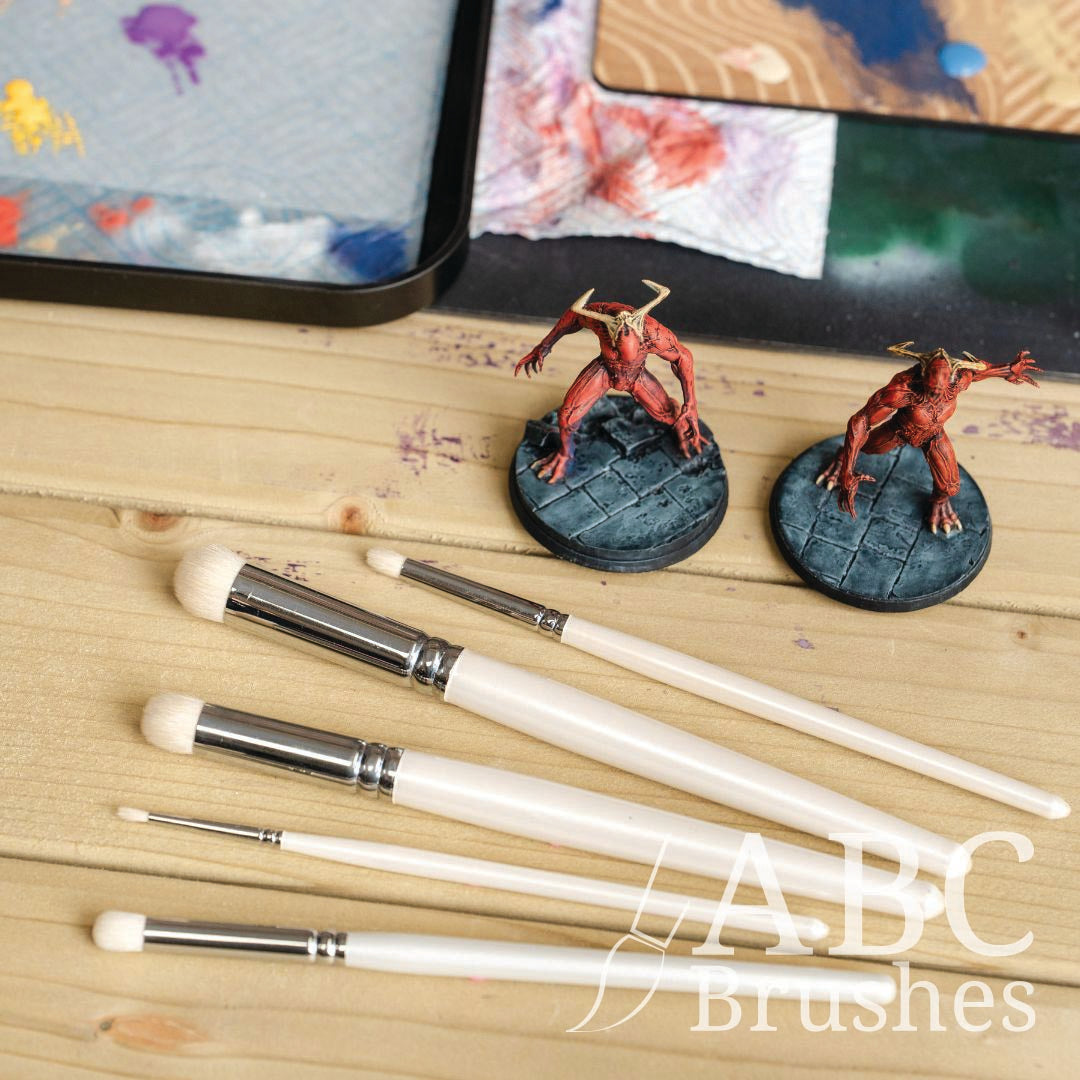 Dry brushes, red demon miniature figures, and a paint palette on a wooden surface.