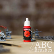 Bottle of the Army Painter Fanatics in the colour Pure Red with various wargaming models and an ABC paintbrush in the background, on a wooden surface.