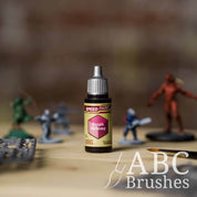 Bottle of the Army Painters Speedpaint in the colour Purple Alchemy, with wargaming models and an ABC paintbrush in the background, on a wooden surface.