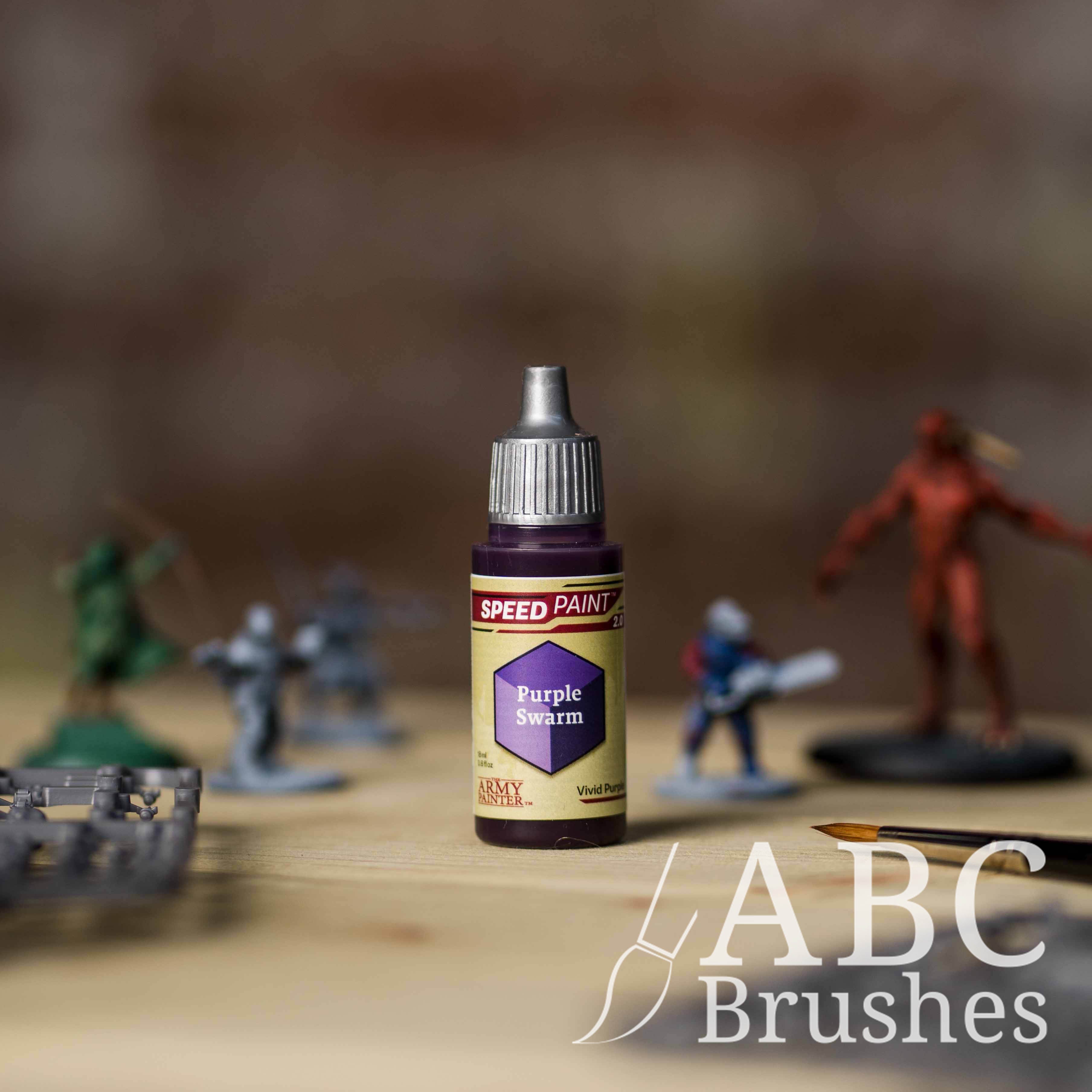 Bottle of the Army Painters Speedpaint in the colour Purple Swarm, with wargaming models and an ABC paintbrush in the background, on a wooden surface.