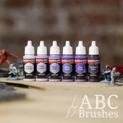 Army Painter Fanatics Purples Triad on a wooden surface. There are wargaming models and an ABC paint brush next to them.