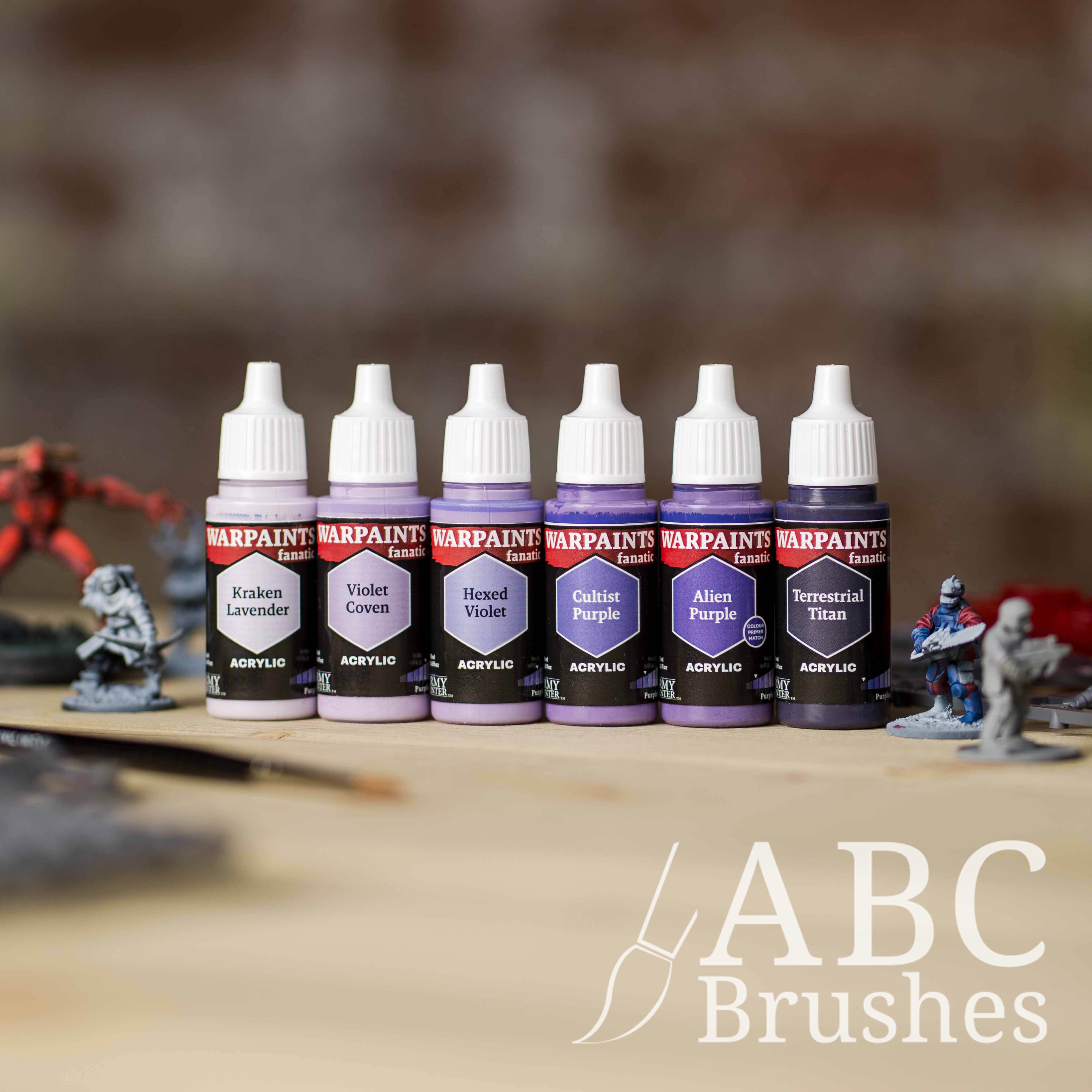Army Painter Fanatics Purples Triad on a wooden surface. There are wargaming models and an ABC paint brush next to them.