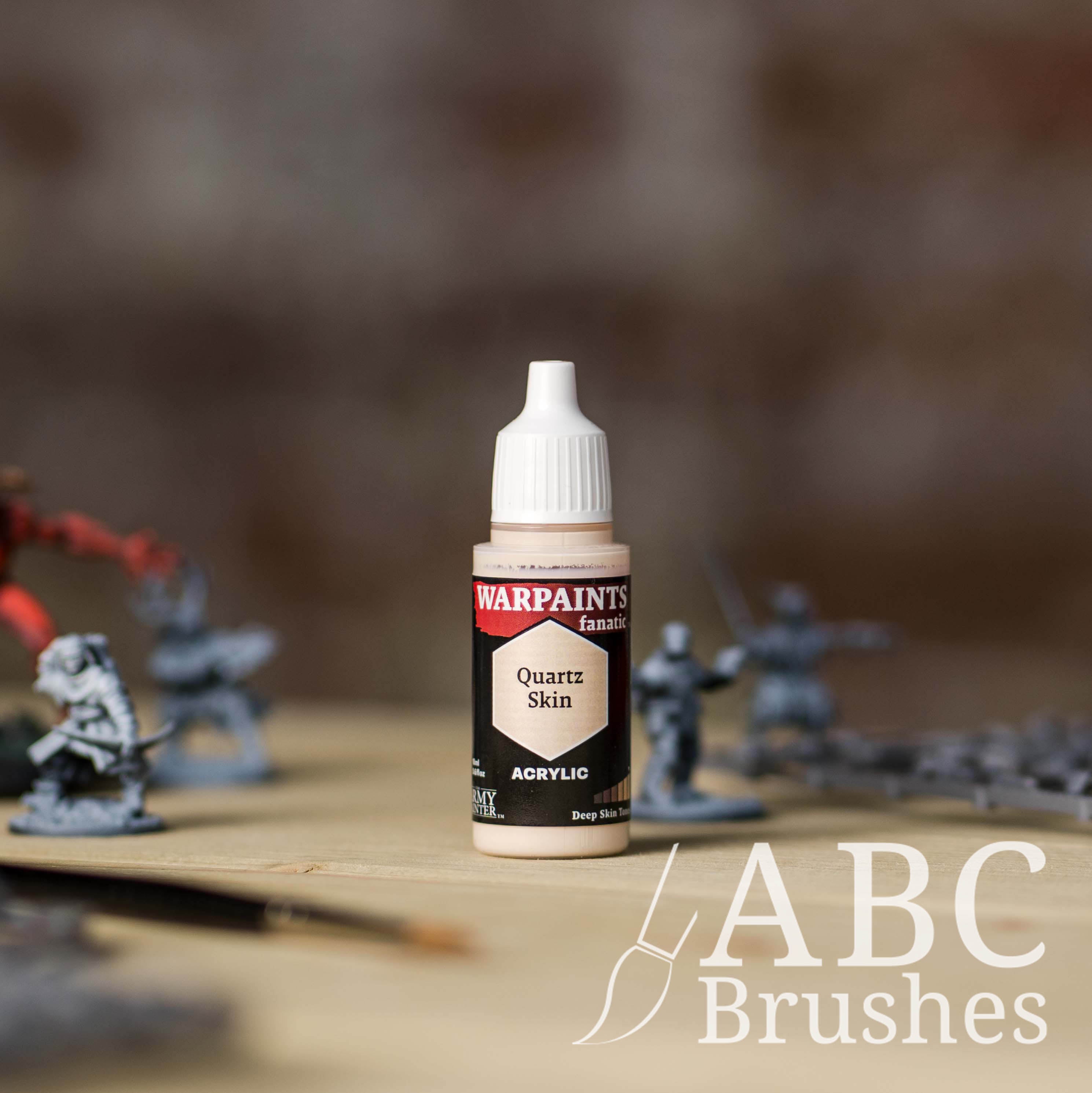 Bottle of the Army Painter Fanatics in the colour Quartz Skin with various wargaming models and an ABC paintbrush in the background, on a wooden surface.
