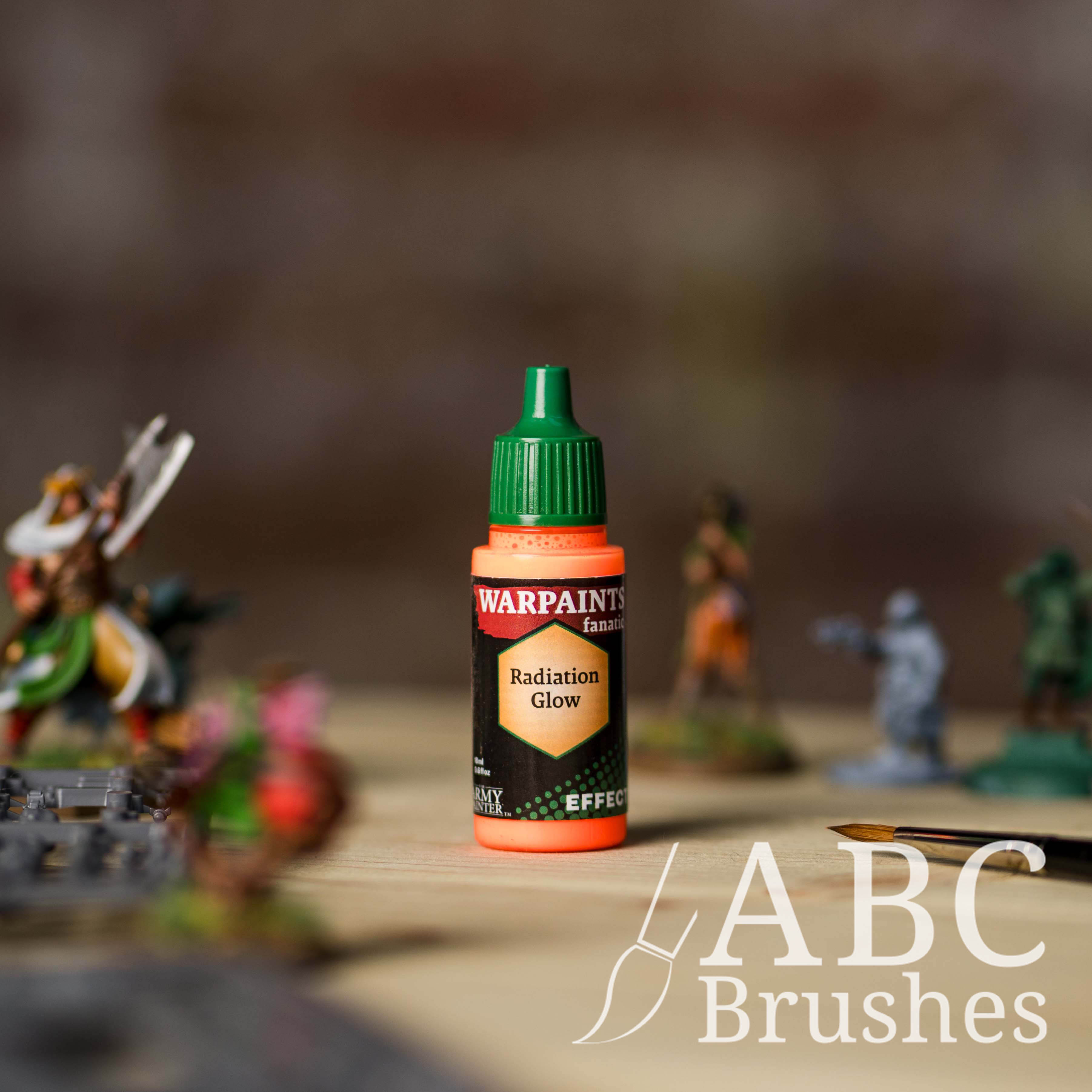 Bottle of the Army Painter Fanatics Effects in the colour Radiation Glow with various wargaming models and an ABC paintbrush in the background, on a wooden surface.