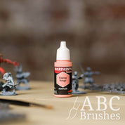 Bottle of the Army Painter Fanatics in the colour Raging Rouge with various wargaming models and an ABC paintbrush in the background, on a wooden surface.
