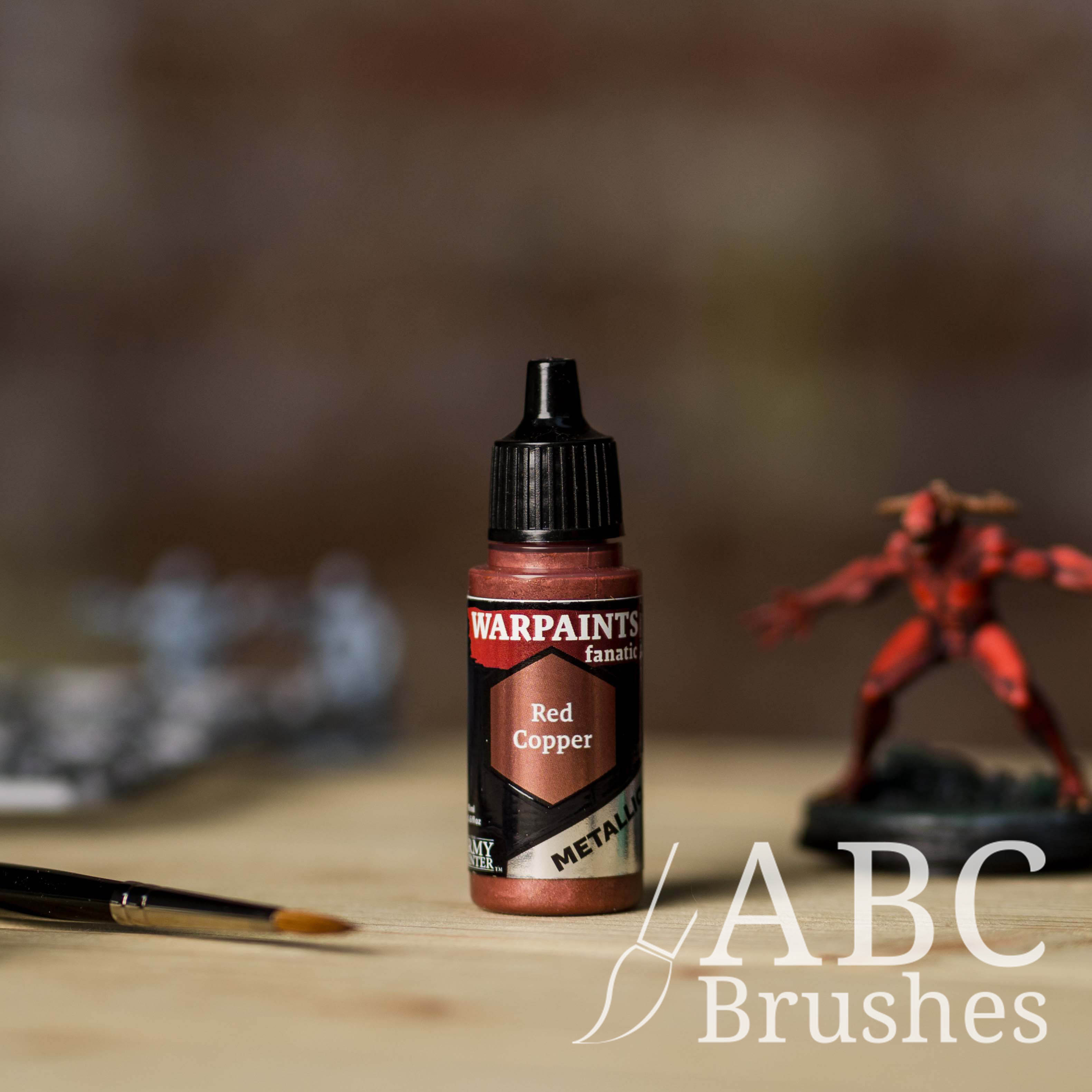 Bottle of the Army Painter Fanatics Metallic paint in the colour Red Copper with various wargaming models and an ABC paintbrush in the background, on a wooden surface.