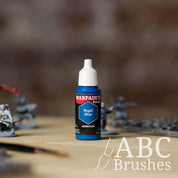 Bottle of the Army Painter Fanatics in the colour Regal Blue with various wargaming models and an ABC paintbrush in the background, on a wooden surface.
