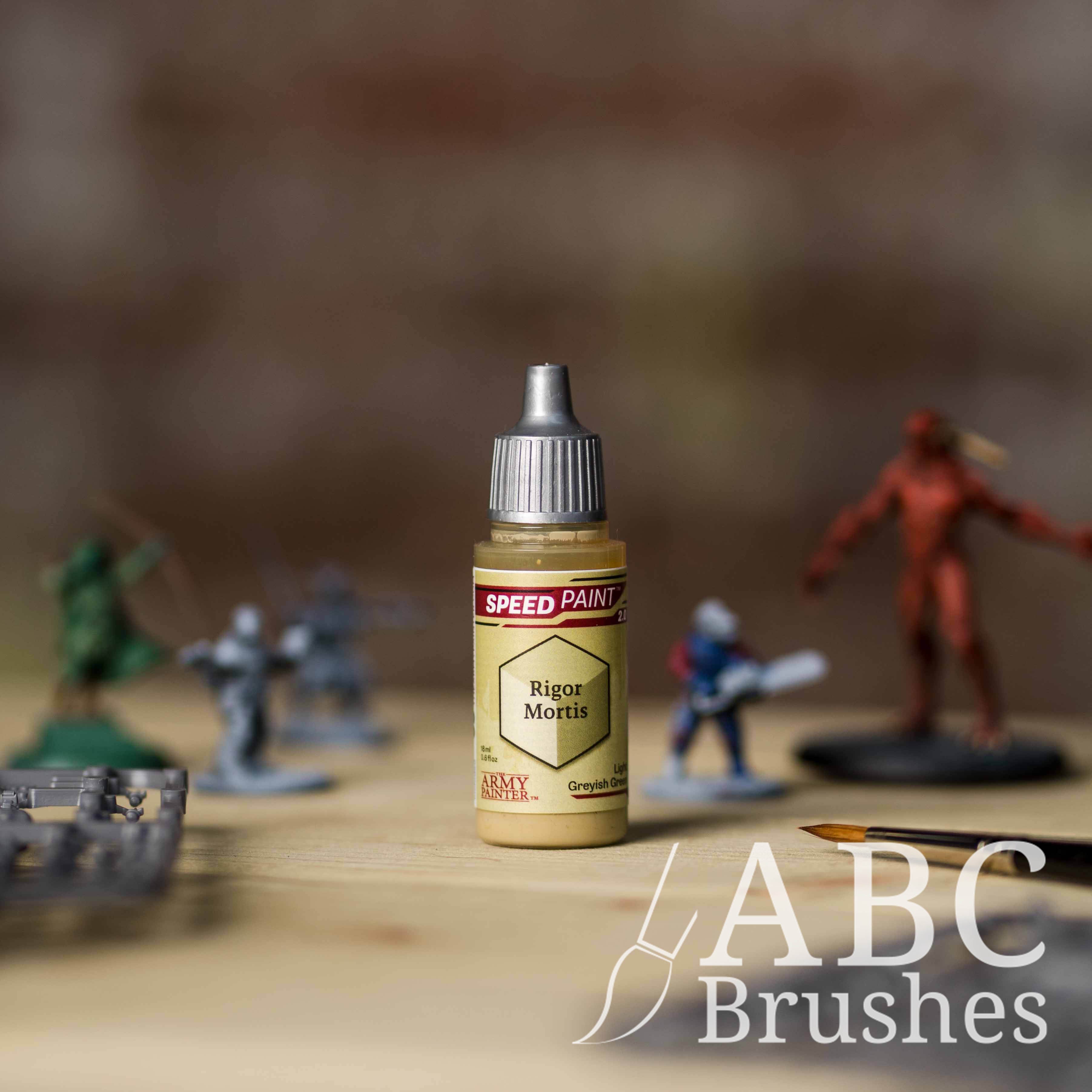 Bottle of the Army Painters Speedpaint in the colour Rigor Mortis, with wargaming models and an ABC paintbrush in the background, on a wooden surface.