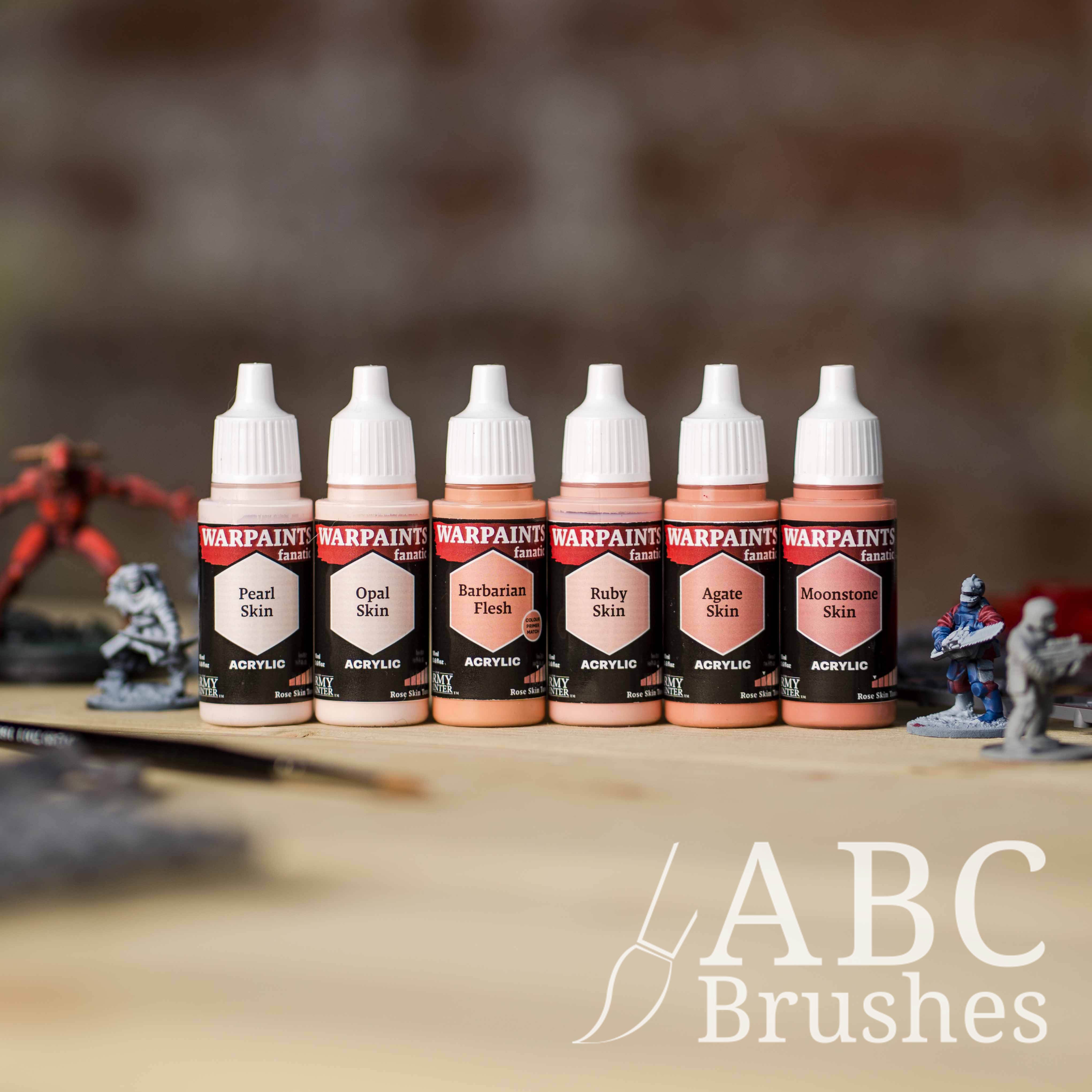 Army Painter Fanatics Rose Skin Tone Triad on a wooden surface. There are wargaming models and an ABC paint brush next to them.