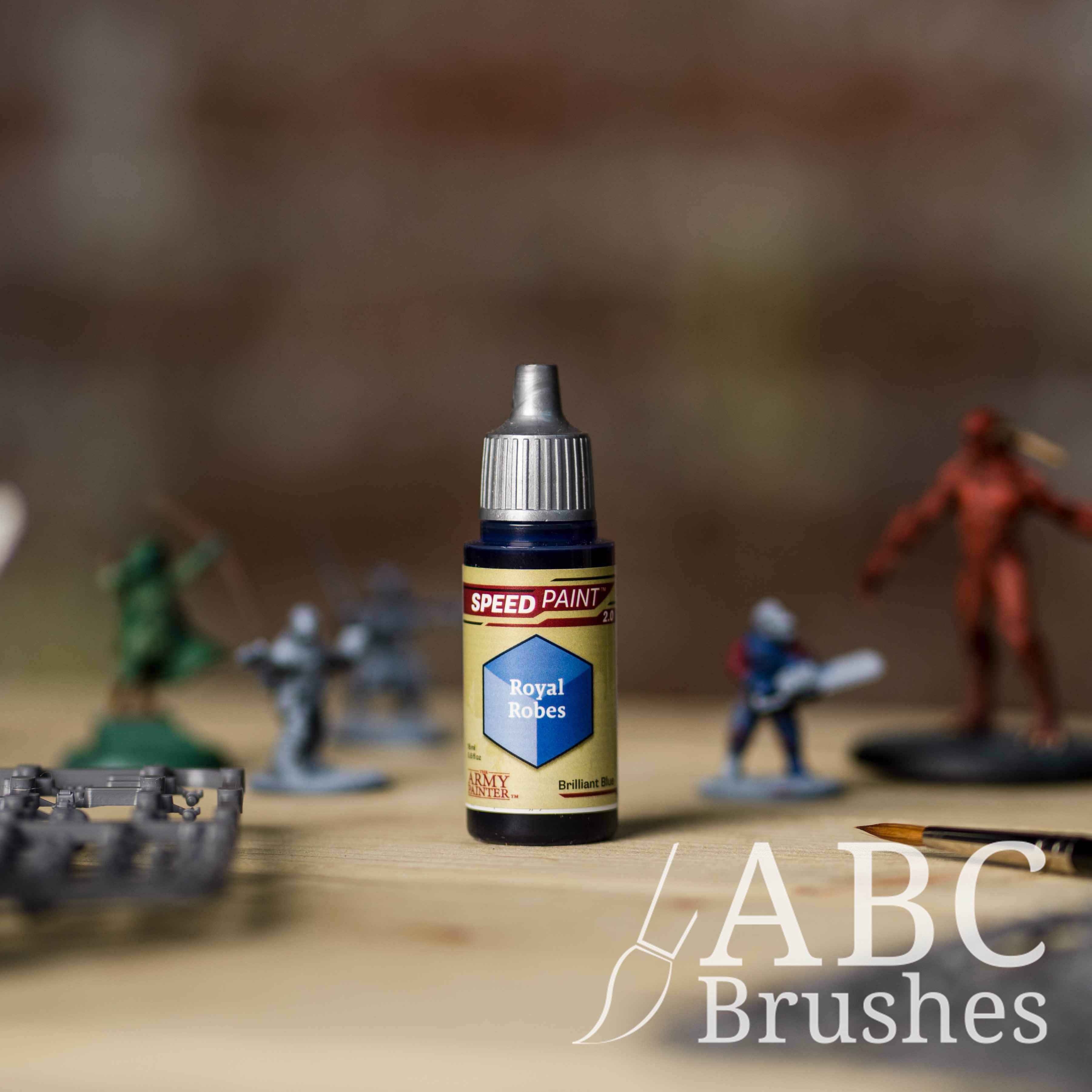 Bottle of the Army Painters Speedpaint in the colour Royal Robes, with wargaming models and an ABC paintbrush in the background, on a wooden surface.