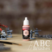Bottle of the Army Painter Fanatics in the colour Ruby Skin with various wargaming models and an ABC paintbrush in the background, on a wooden surface.