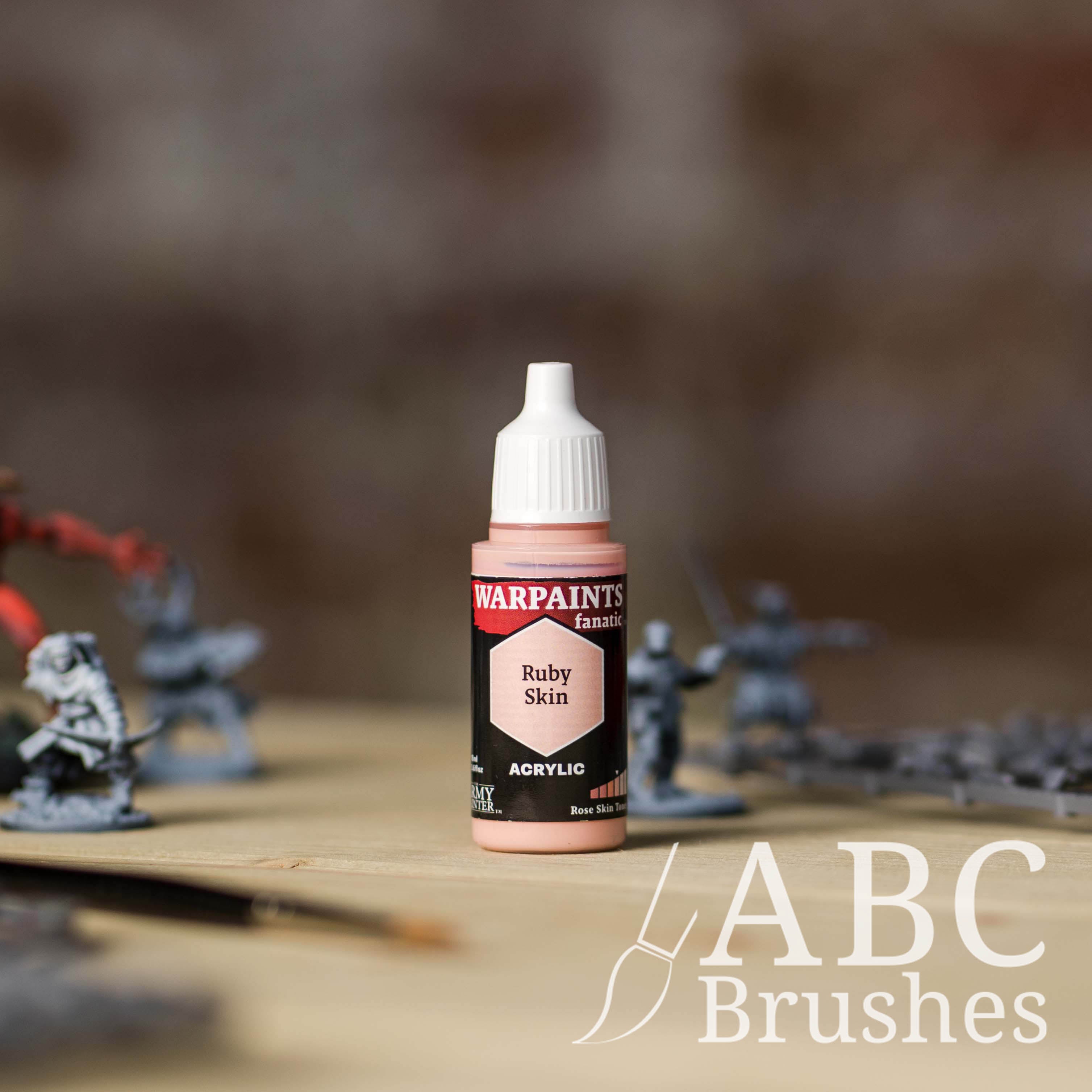 Bottle of the Army Painter Fanatics in the colour Ruby Skin with various wargaming models and an ABC paintbrush in the background, on a wooden surface.