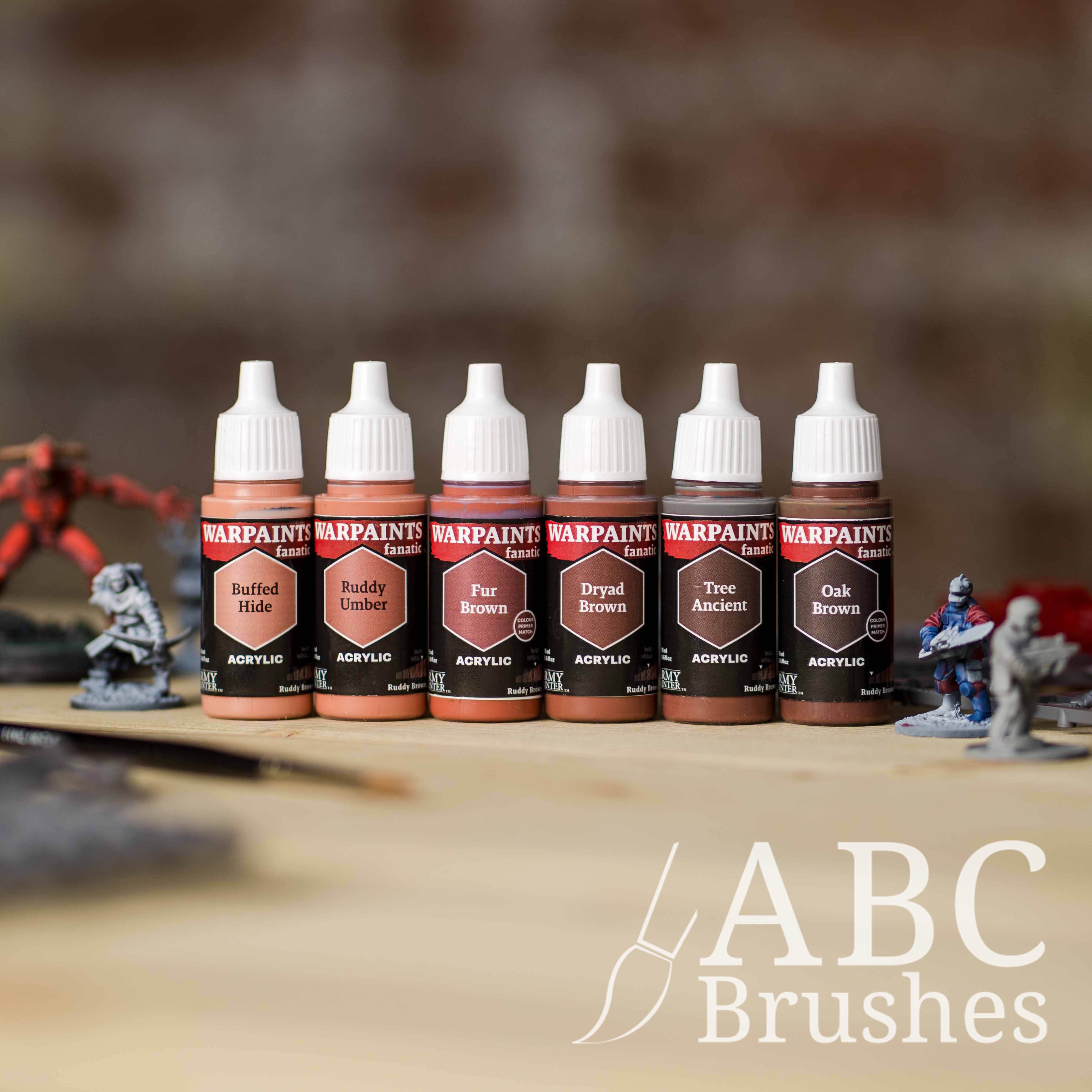 Army Painter Fanatics Ruddy Browns Triad on a wooden surface. There are wargaming models and an ABC paint brush next to them.