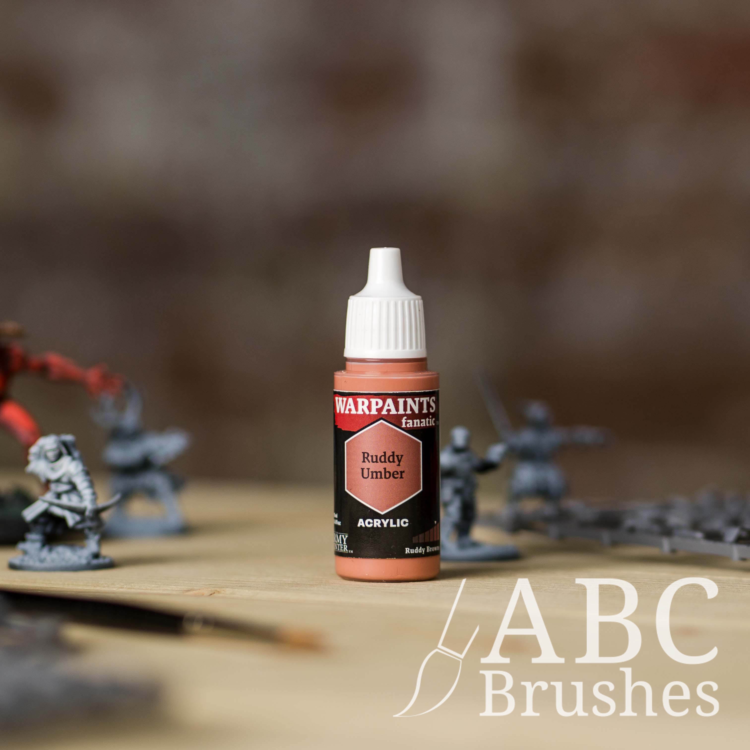 Bottle of the Army Painter Fanatics in the colour Ruddy Umber with various wargaming models and an ABC paintbrush in the background, on a wooden surface.