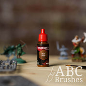 Bottle of the Army Painter Fanatics Wash in the colour Rust Tone with various wargaming models and an ABC paintbrush in the background, on a wooden surface.