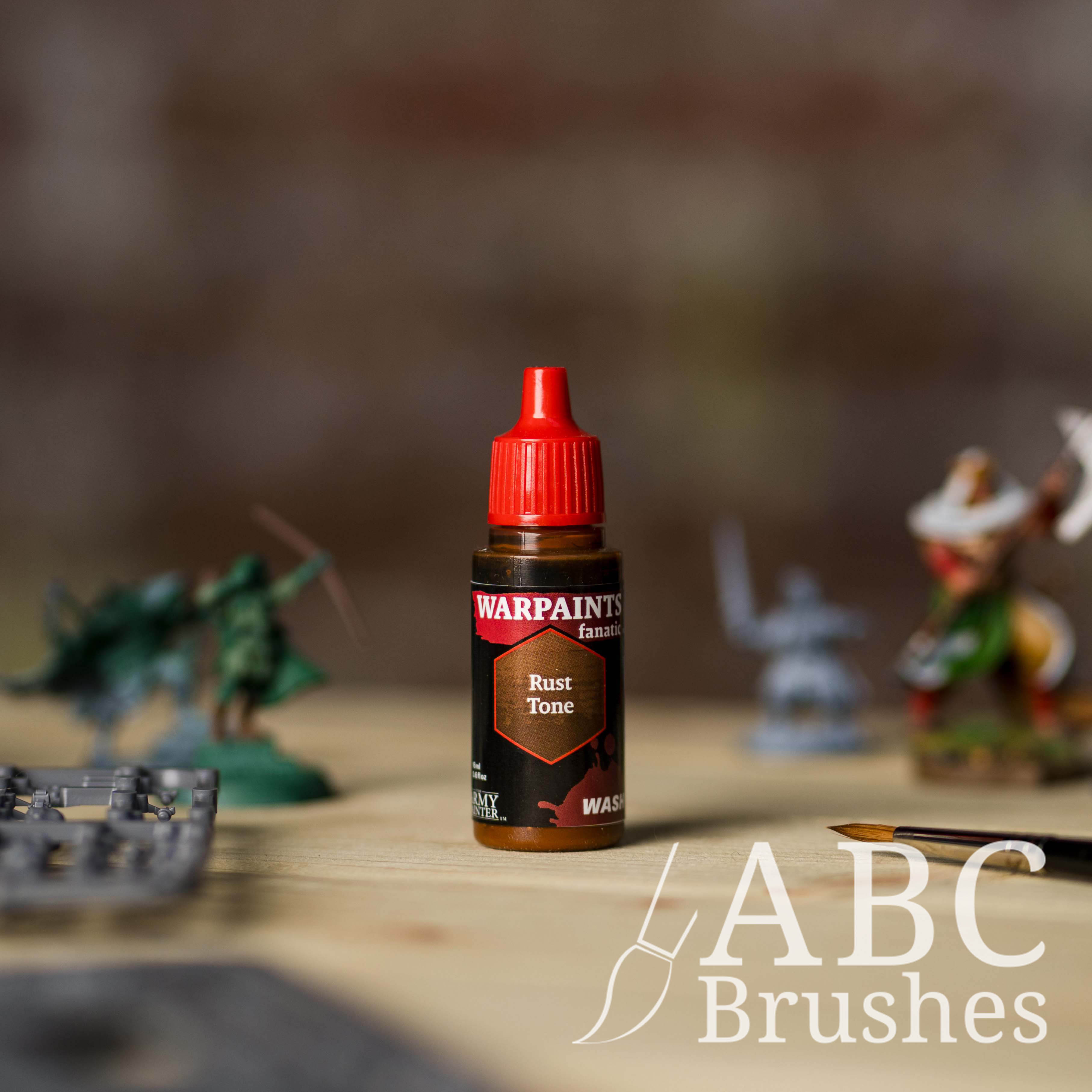 Bottle of the Army Painter Fanatics Wash in the colour Rust Tone with various wargaming models and an ABC paintbrush in the background, on a wooden surface.