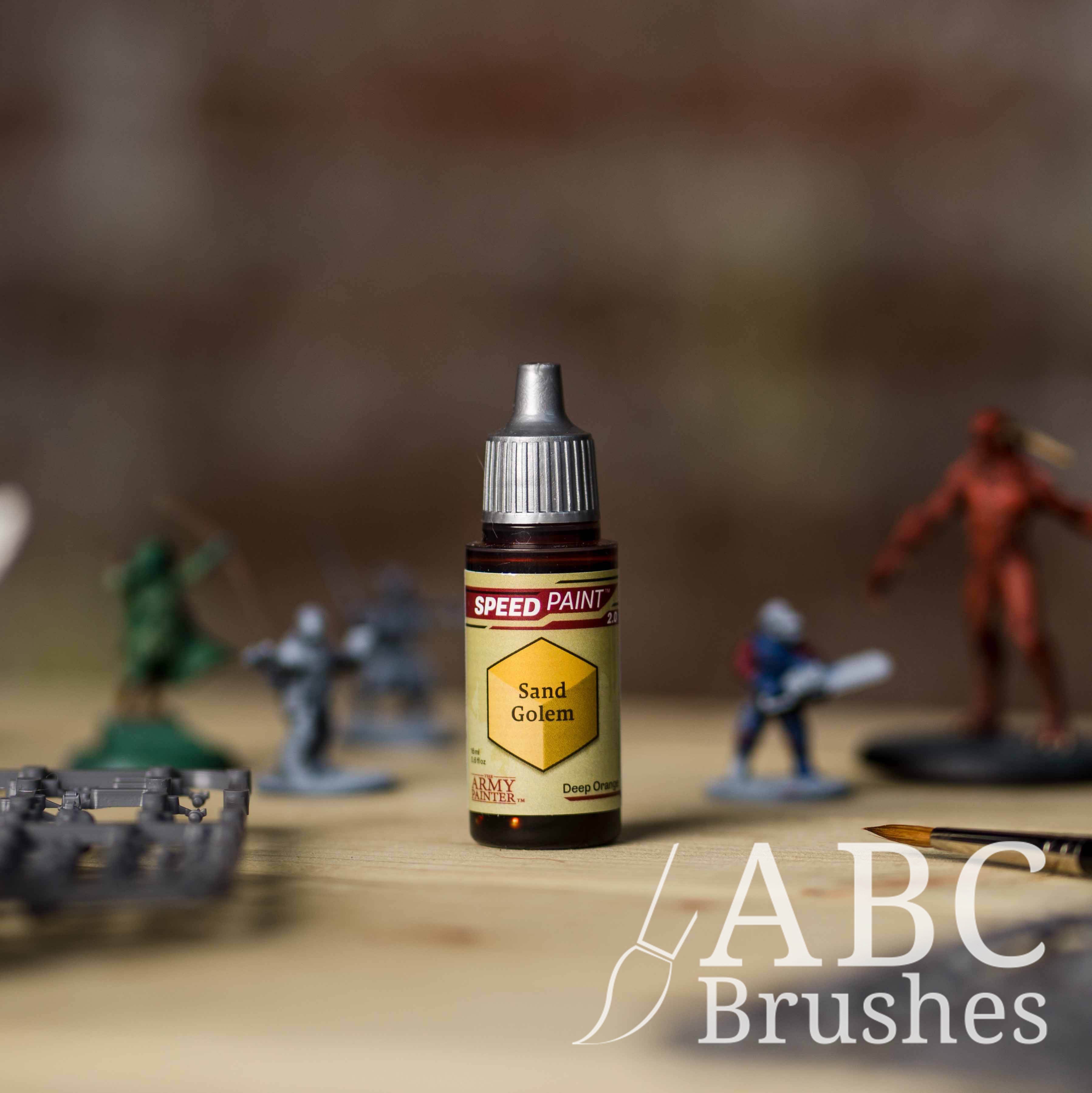 Bottle of the Army Painters Speedpaint in the colour Sand Golem, with wargaming models and an ABC paintbrush in the background, on a wooden surface.
