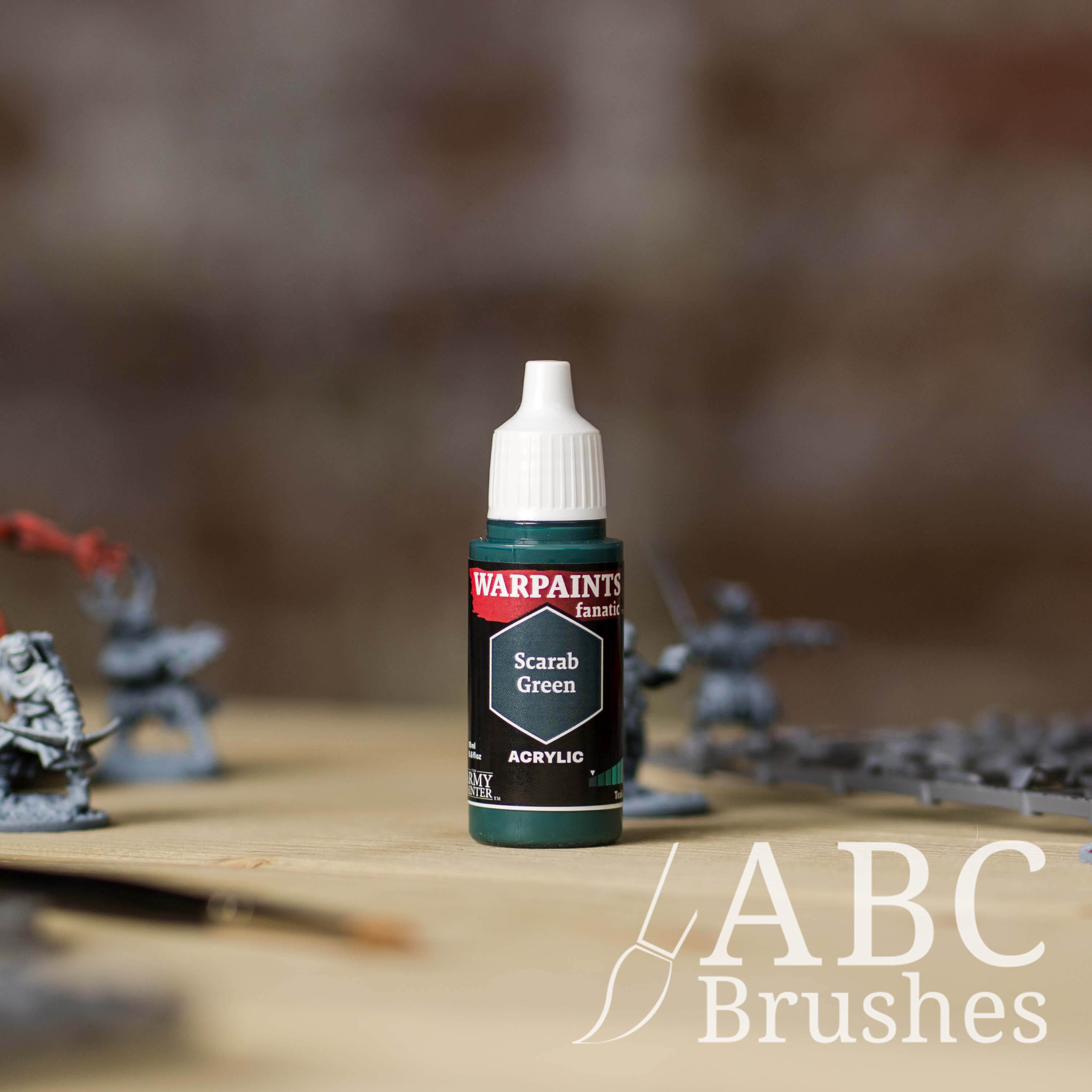 Bottle of the Army Painter Fanatics in the colour Scarab Green with various wargaming models and an ABC paintbrush in the background, on a wooden surface.