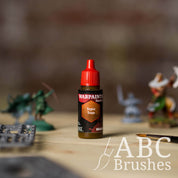 Bottle of the Army Painter Fanatics Wash in the colour Sepia Tone with various wargaming models and an ABC paintbrush in the background, on a wooden surface.