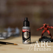 Bottle of the Army Painter Fanatics Metallic paint in the colour Shining Silver with various wargaming models and an ABC paintbrush in the background, on a wooden surface.