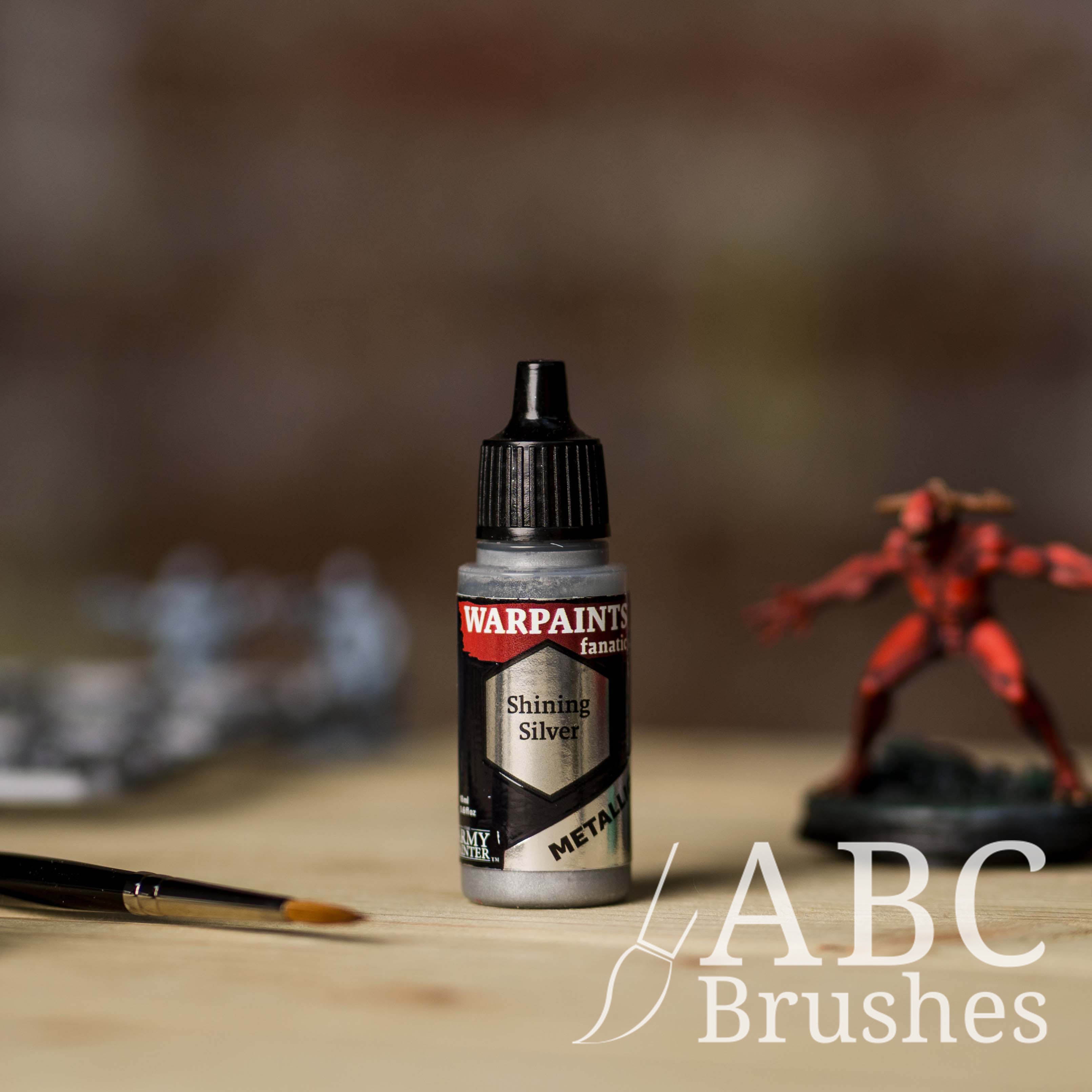 Bottle of the Army Painter Fanatics Metallic paint in the colour Shining Silver with various wargaming models and an ABC paintbrush in the background, on a wooden surface.