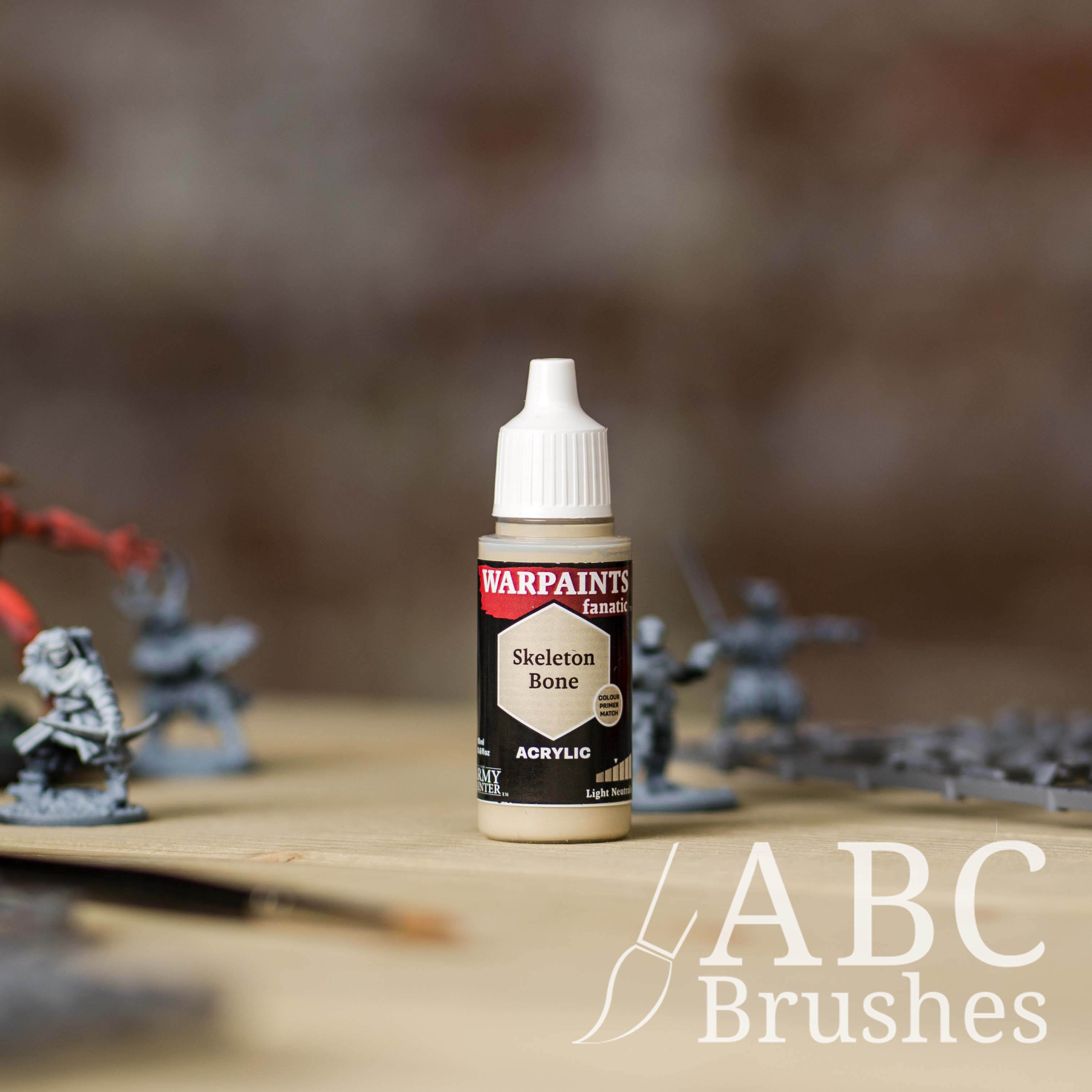 Bottle of the Army Painter Fanatics in the colour Skeleton Bone with various wargaming models and an ABC paintbrush in the background, on a wooden surface.