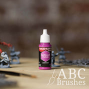 Bottle of the Army Painter Fanatics in the colour Spellbound Fuchsia with various wargaming models and an ABC paintbrush in the background, on a wooden surface.