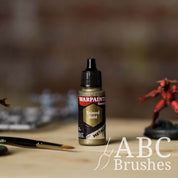 Bottle of the Army Painter Fanatics Metallic paint in the colour Tainted Gold with various wargaming models and an ABC paintbrush in the background, on a wooden surface.