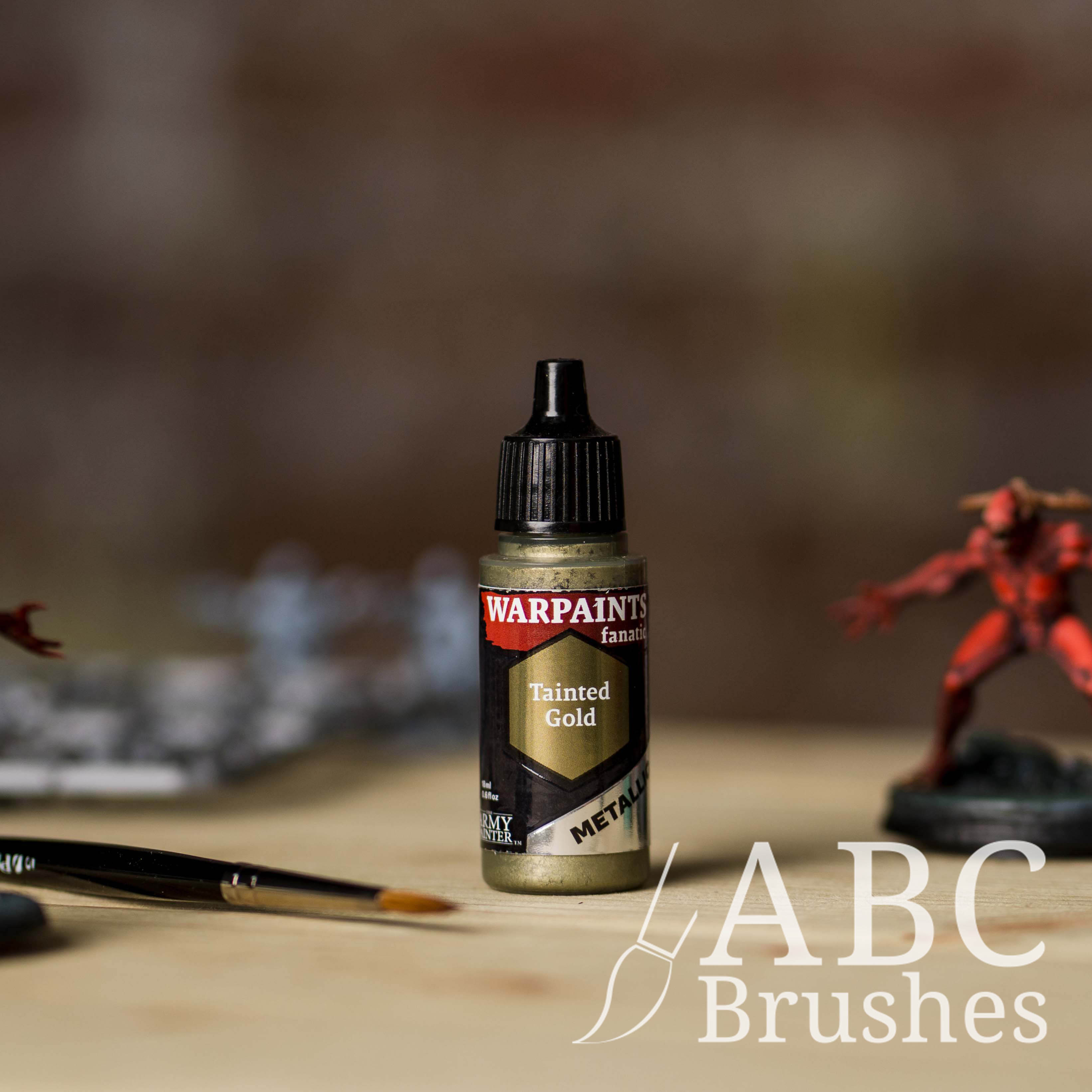 Bottle of the Army Painter Fanatics Metallic paint in the colour Tainted Gold with various wargaming models and an ABC paintbrush in the background, on a wooden surface.