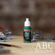 Bottle of the Army Painter Fanatics in the colour Temple Gate Teal with various wargaming models and an ABC paintbrush in the background, on a wooden surface.