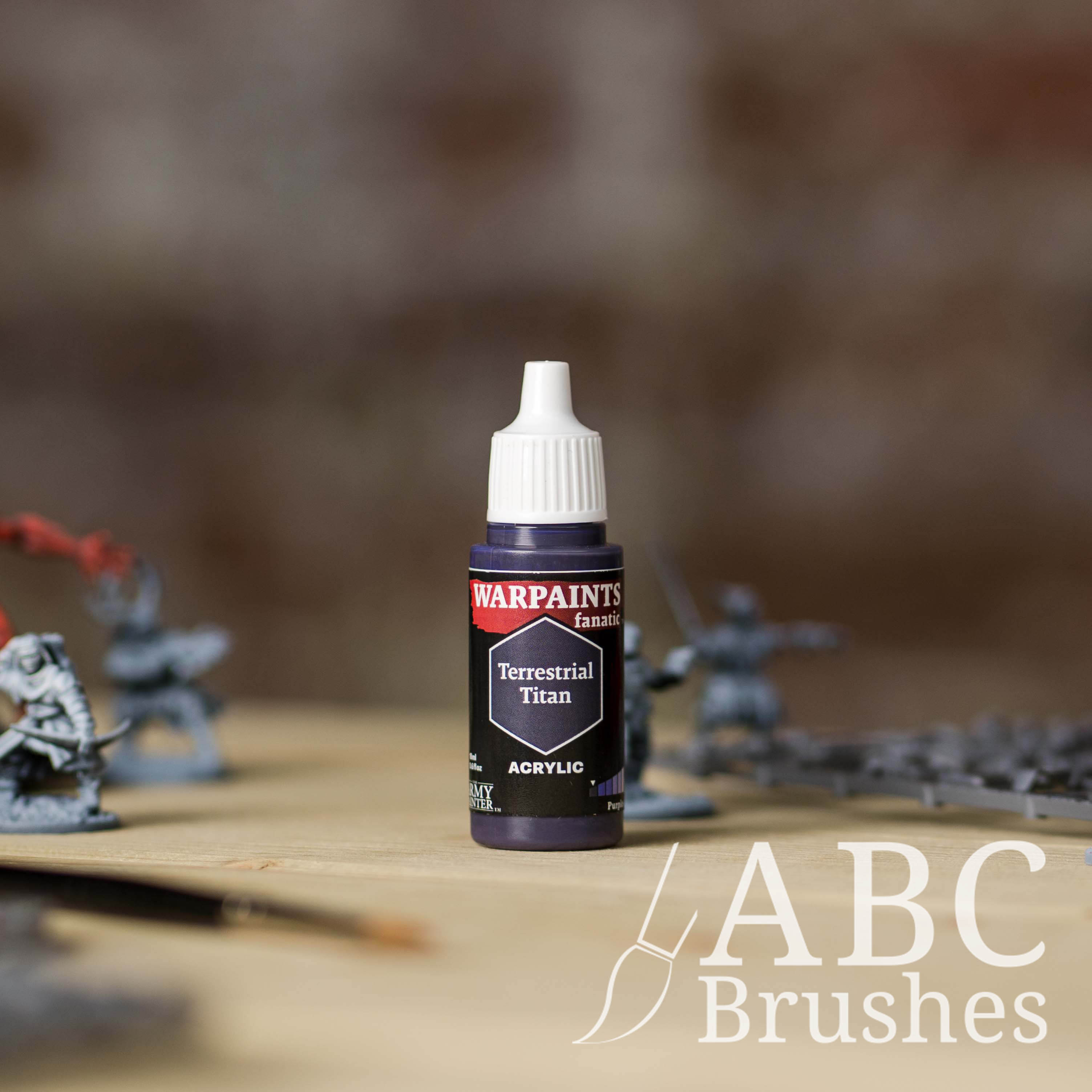 Bottle of the Army Painter Fanatics in the colour Terrestrial Titan with various wargaming models and an ABC paintbrush in the background, on a wooden surface.