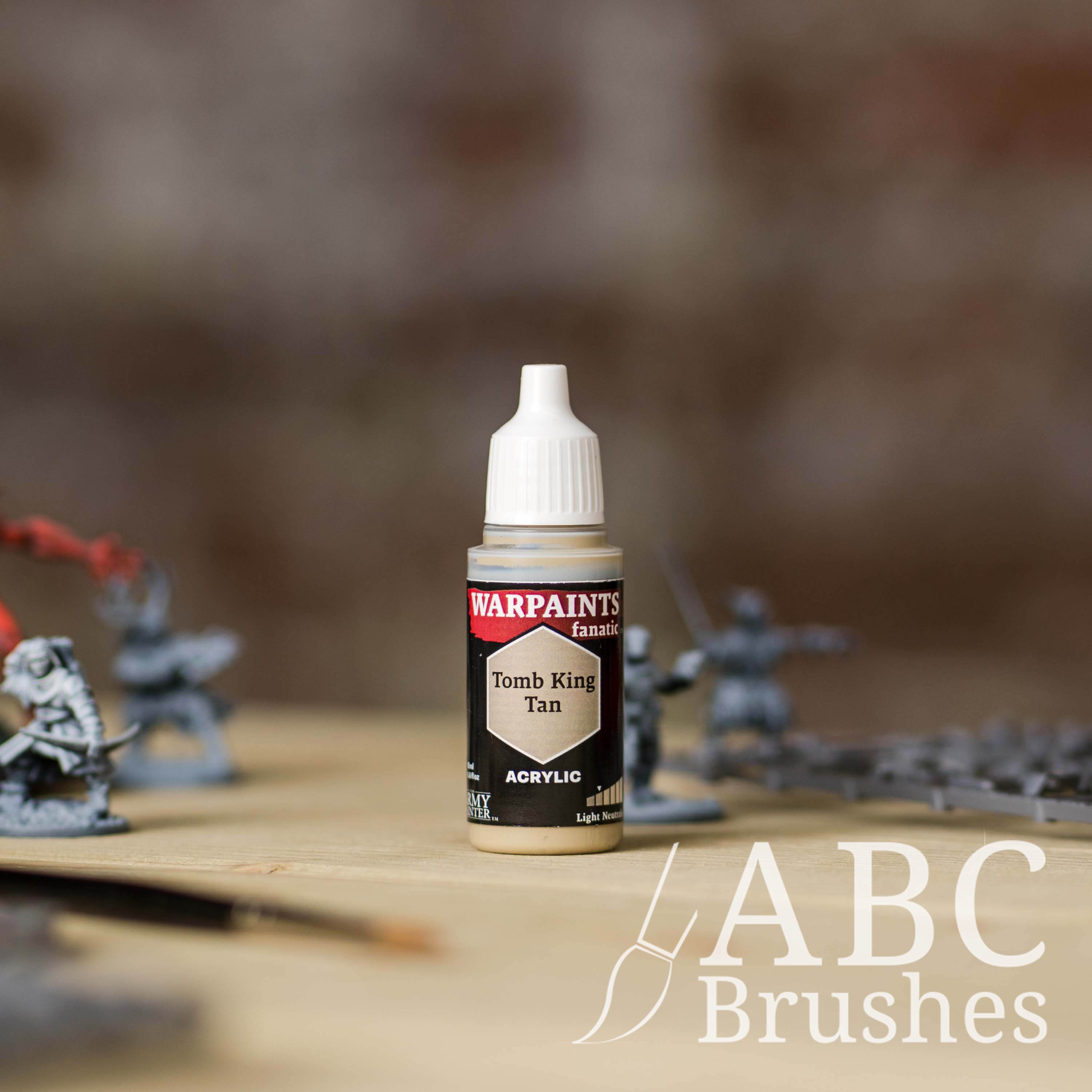 Bottle of the Army Painter Fanatics in the colour Tomb King Tan with various wargaming models and an ABC paintbrush in the background, on a wooden surface.