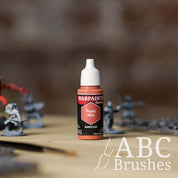 Bottle of the Army Painter Fanatics in the colour Topaz Skin with various wargaming models and an ABC paintbrush in the background, on a wooden surface.