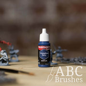 Bottle of the Army Painter Fanatics in the colour Triumphant Navy with various wargaming models and an ABC paintbrush in the background, on a wooden surface.