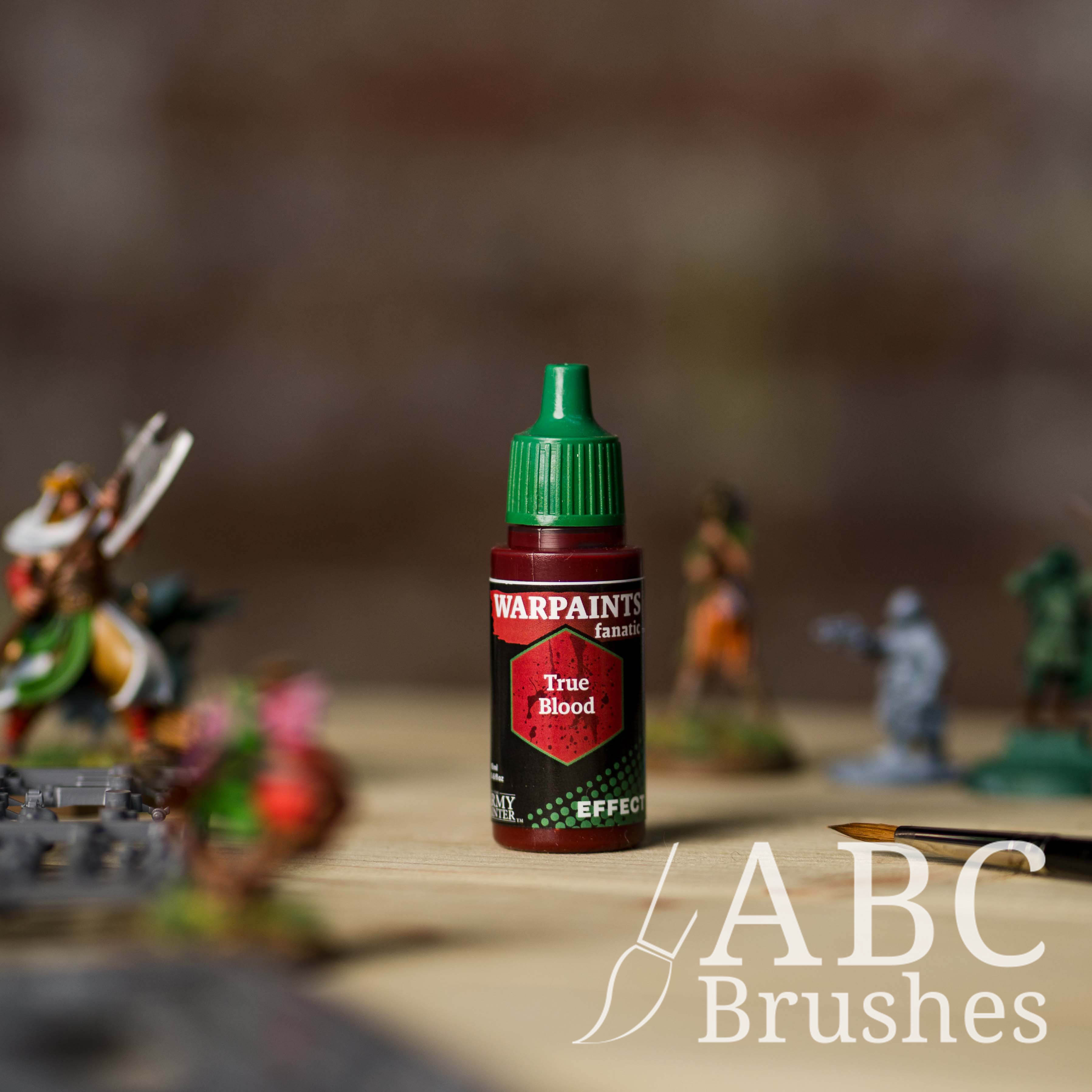 Bottle of the Army Painter Fanatics Effects in the colour True Blood with various wargaming models and an ABC paintbrush in the background, on a wooden surface.