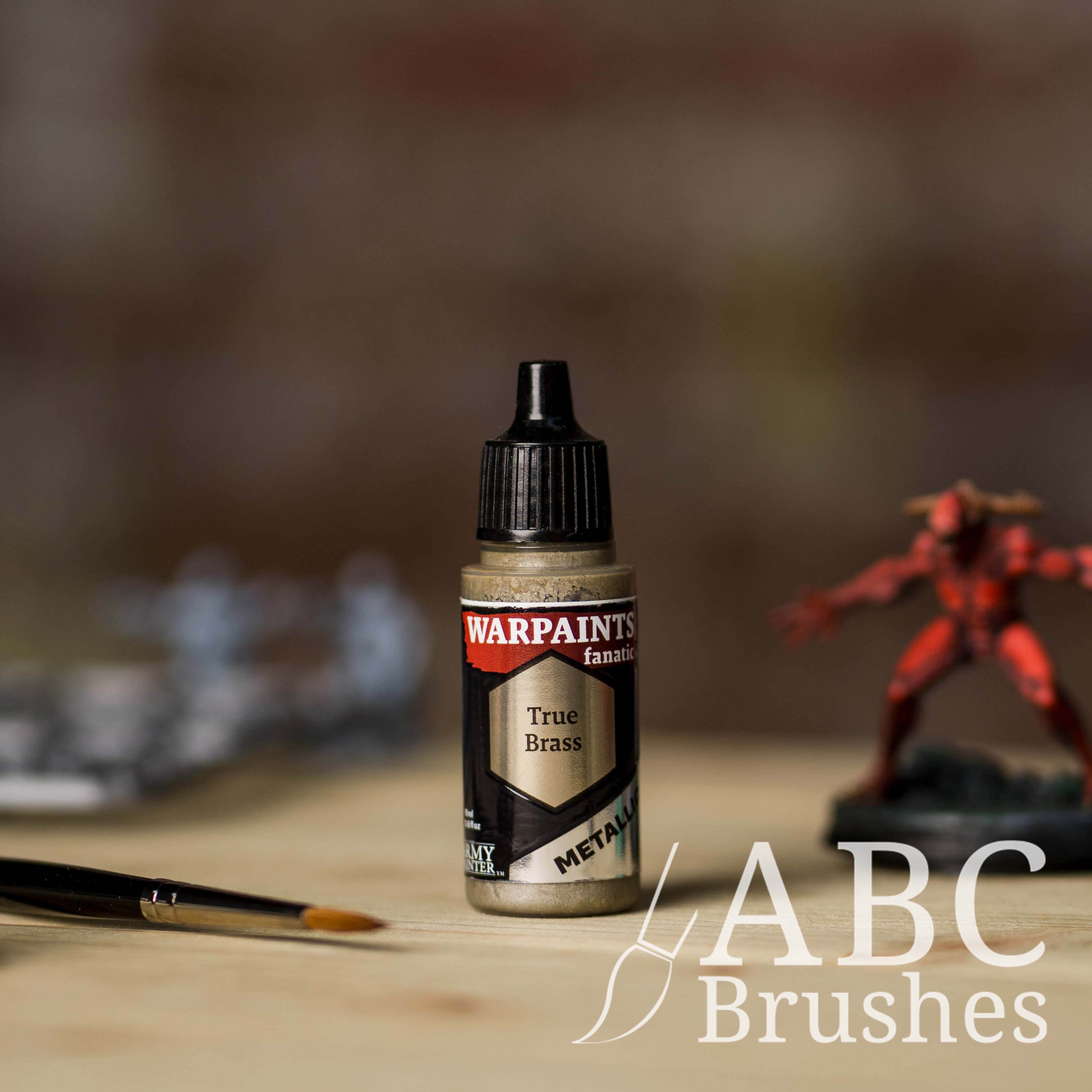 Bottle of the Army Painter Fanatics Metallic paint in the colour True Brass with various wargaming models and an ABC paintbrush in the background, on a wooden surface.