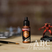Bottle of the Army Painter Fanatics Metallic paint in the colour True Copper with various wargaming models and an ABC paintbrush in the background, on a wooden surface.