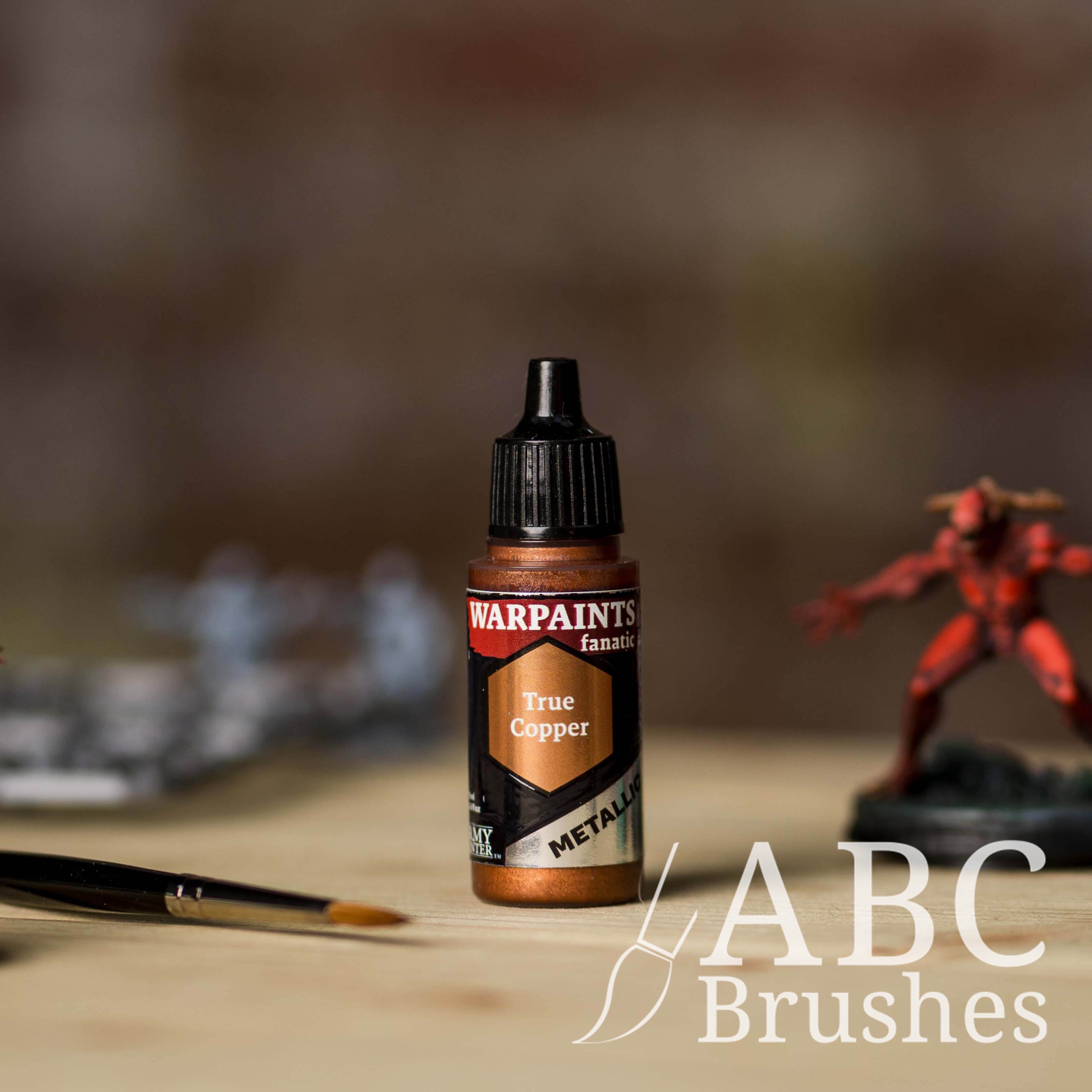 Bottle of the Army Painter Fanatics Metallic paint in the colour True Copper with various wargaming models and an ABC paintbrush in the background, on a wooden surface.