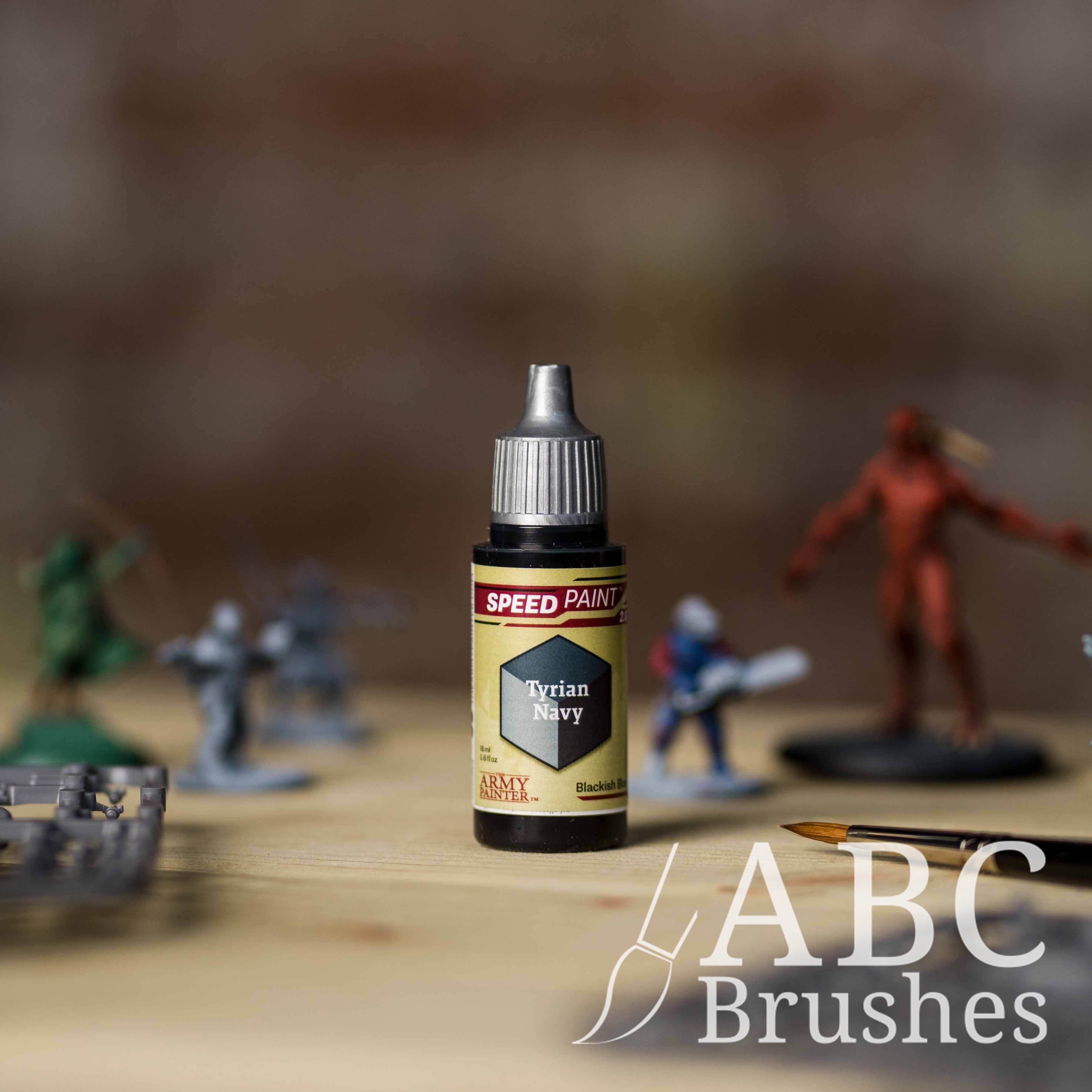 Bottle of the Army Painters Speedpaint in the colour Tyrian Navy, with wargaming models and an ABC paintbrush in the background, on a wooden surface.