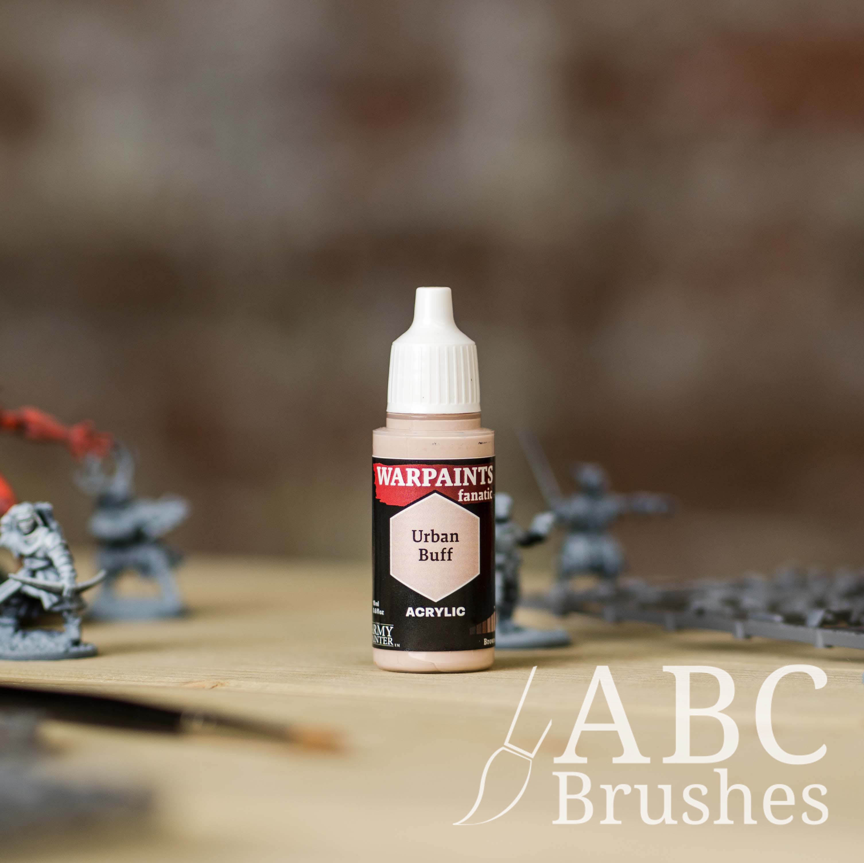 Bottle of the Army Painter Fanatics in the colour Urban Buff with various wargaming models and an ABC paintbrush in the background, on a wooden surface.