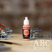 Bottle of the Army Painter Fanatics in the colour Violent Vermillion with various wargaming models and an ABC paintbrush in the background, on a wooden surface.